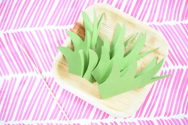 Paper pineapple stems folded in half lay in a square paper faux-wooden craft plate.
