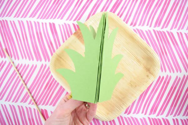 Above a background of pink slashes on white paper, Abbi Kirsten's hand holds a pineapple stem made of four pieces of green paper. After assembly the green paper will make a three-dimensional pineapple crown. 