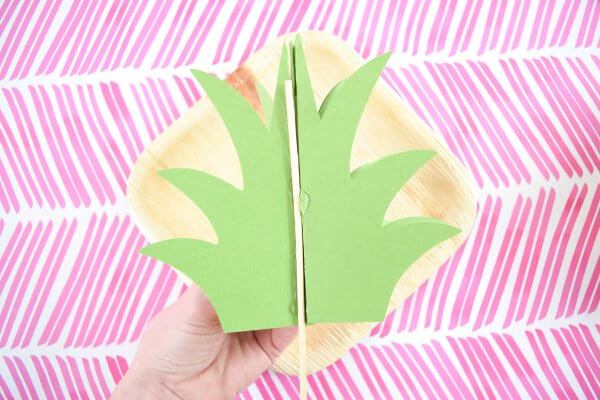 Above a white and pink background, Abbi Kirsten's hand demonstrates how to glue a bamboo skewer into the middle of a paper pineapple crown.