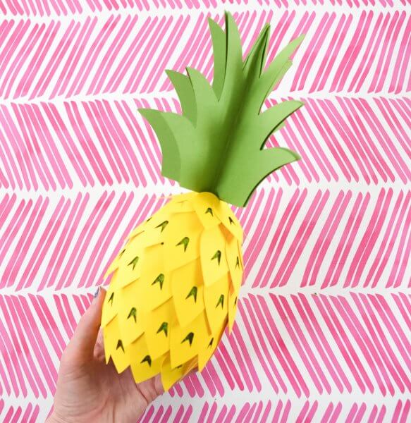 A paper pineapple party decoration craft is held by Abbi Kirsten's hand over a pink and white festive background. 