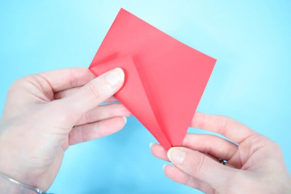 A woman holds a red paper square in her hand and folds one edge of the square inwards.
