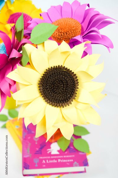 A bouquet of colorful paper sunflowers; pink, red, and purple sunflowers sit on top of two stacked books.