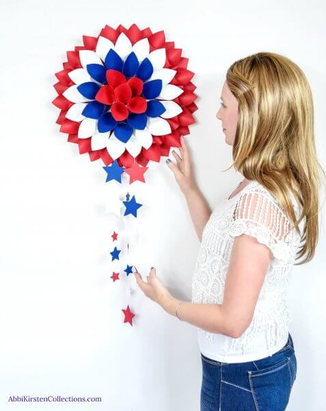 A tall blonde woman wearing a white shirt poses with a red, white, and blue wreath hanging on the wall.