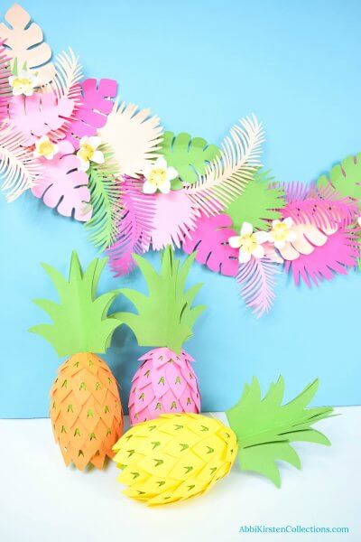 Three DIY pineapple party decorations sit on a white surface against a light blue wall. Behind the pineapples is a paper banner of tropical luau party flowers. "AbbiKirstenCollections.com" is typed in the bottom right corner. 