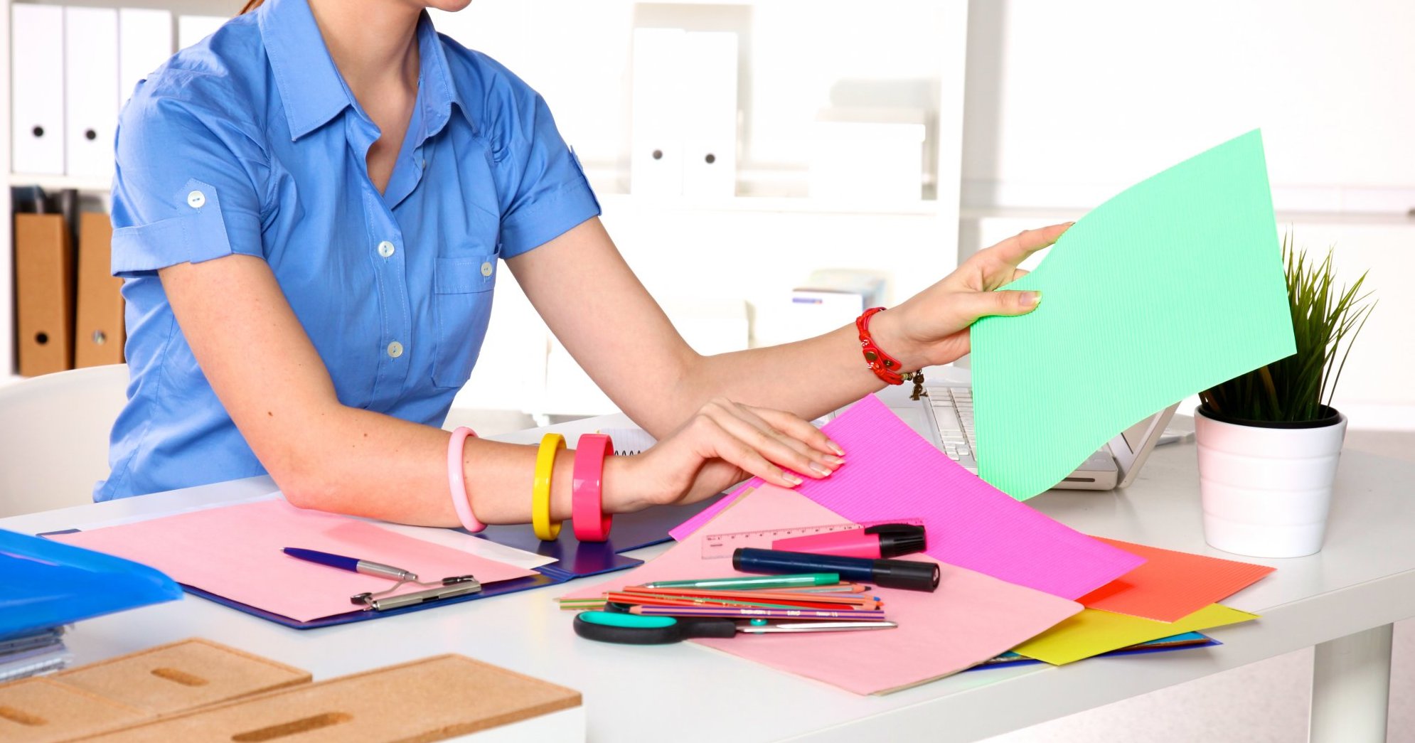 A woman wearing a short-sleeved blue button-down shirt sits at a desk, arranging sheets of paper in various colors. Other craft supplies like markers, rulers, and a scissors sit on the desk.