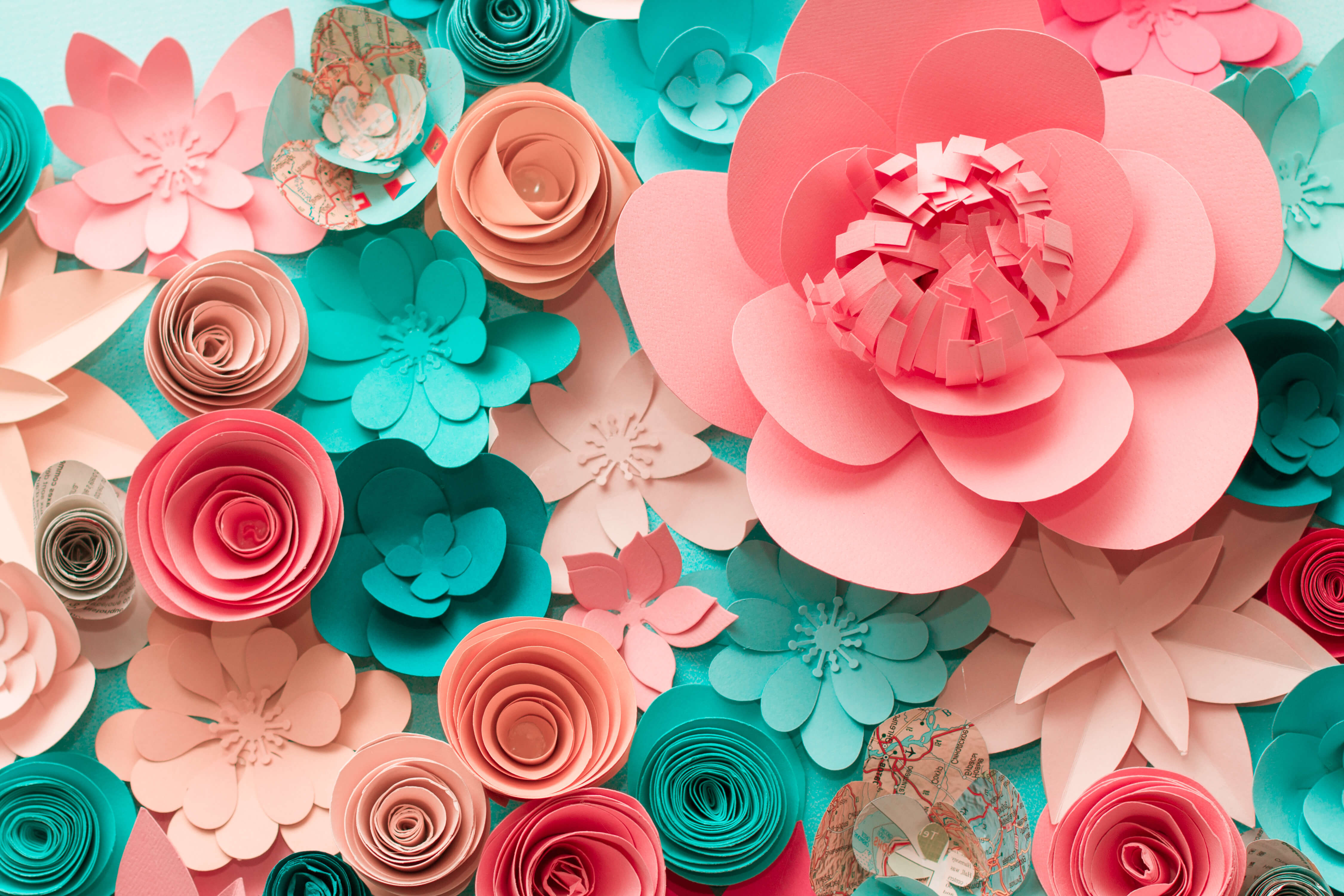 A collection of many different paper flowers in different styles. The pink, coral, and teal colored paper flowers are arranged on a table, and the image is an overhead angle looking down.