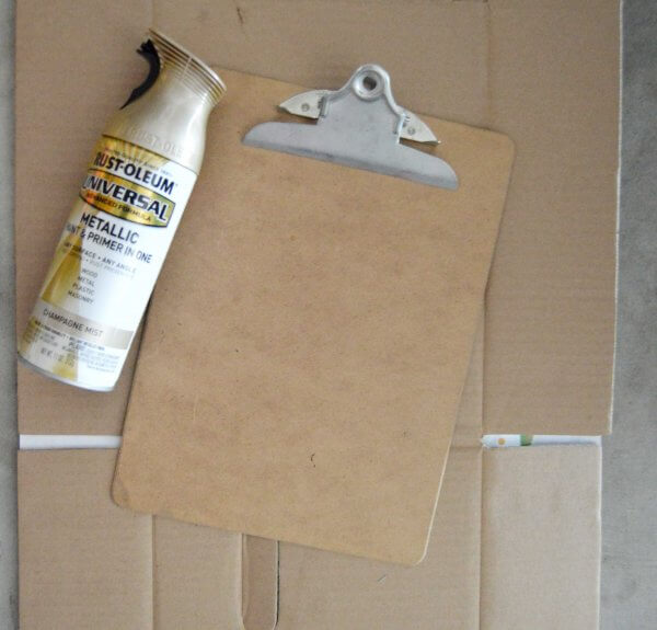 A can of Rustoleum metallic spray lays next to a clipboard on top of cardboard, ready to be painted. 