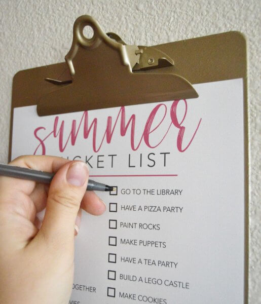 A close-up of a woman's hand holding a pen while she checks off "go to the library," an item on a summer bucket checklist. 