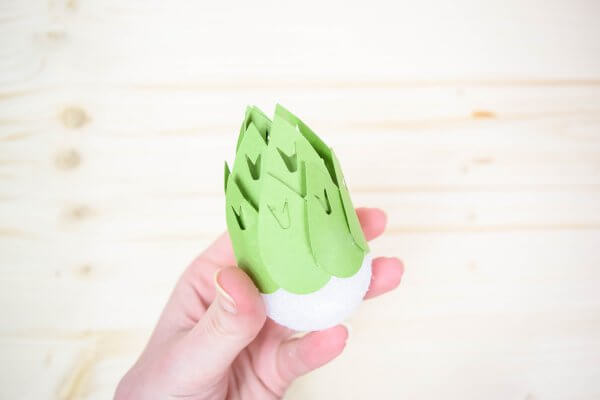 A hand holding a white foam ball with pieces of light green paper attached forming the stamen of a magnolia. 