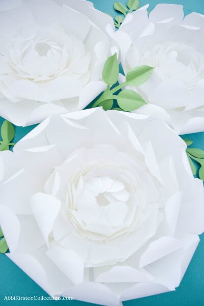 A closeup of the center of a giant snowy paper peony flower with two more peonies in the background. The green leaves are a stark contrast to the snowy white, delicate petals of the craft flowers. 