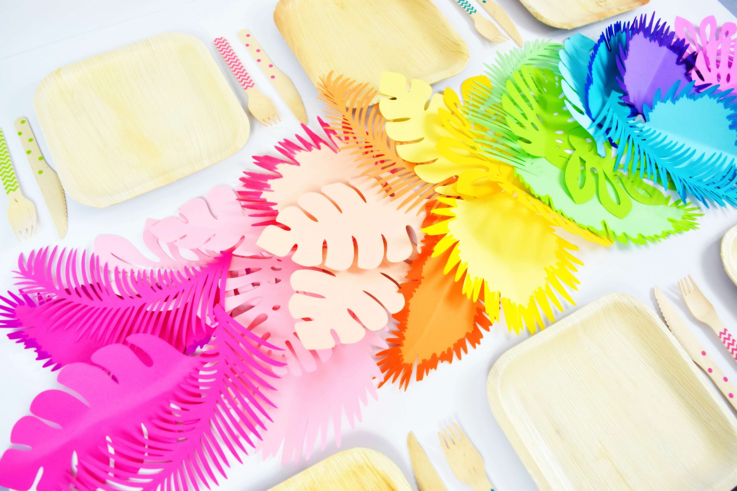 A tropical rainbow colored centerpiece runs down a table set with bamboo plate place settings. The table runner us made up of large tropical style leaves.