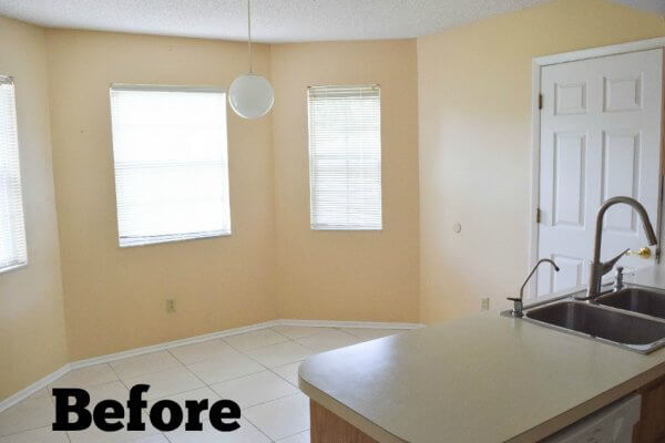 A different view of an outdated beige, empty kitchen shows a small in-kitchen dining area in the shape of a half-octagon. A lone round white ball-shaped light fixture hangs over the empty space. 