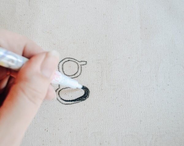 A hand holding a black fabric marker coloring in a lowercase "g" on cream fabric.