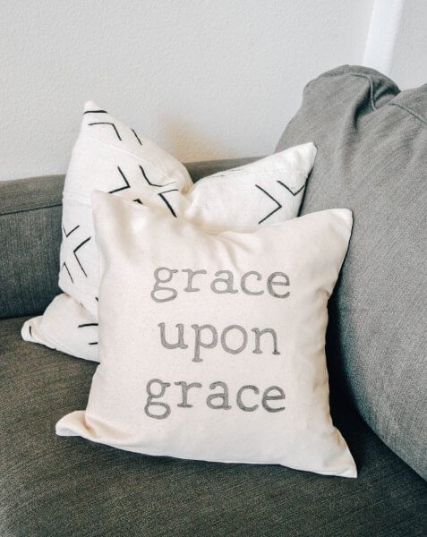 Two cream colored pillows on a grey couch. One pillow has "grace upon grace" written on it.  