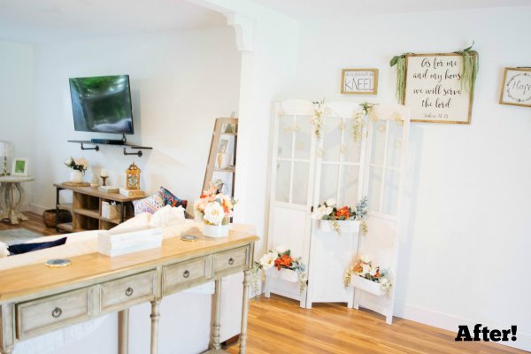 The family room is decorated with white furniture and a light-wood table behind the couch. A decorative white screen full of paper craft flowers is in the corner, and craft prints adorn the white walls. TV and other wooden consoles and end tables are also visble. The word "After! is in the lower right corner. This room is the after photo of a renovation of Abbi Kirsten's home. 