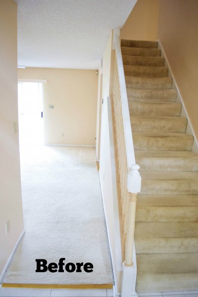 A dirty yellow staircase goes to the second floor of a house in desperate need of a renovation. Next to the stairway is an open hallway to a well-lit back room. 