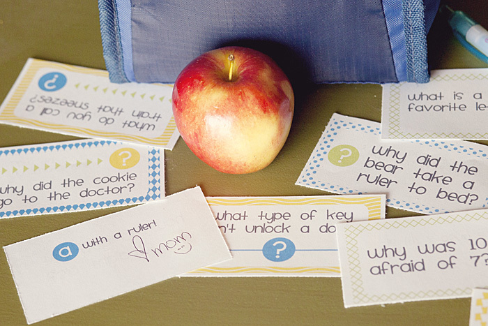 Small paper lunch box notes with jokes and brain teasers lay spread out around a child's lunch box, with a fresh apple in the center.