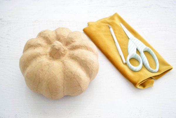 A craft pumpkin, a length of folded gold velvet fabric, scissors, and an Exacto knife are laid out on a table. These supplies are used to make no-sew fabric pumpkins. 