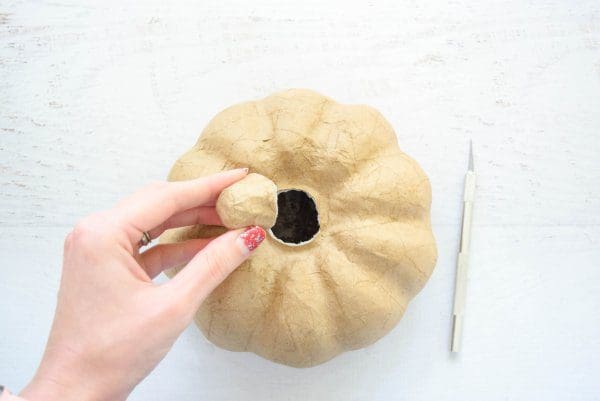 Abbi removes the stem from the paper mache pumpkin, leaving a hole at the top. An Exacto knife lies to the right of the pumpkin.