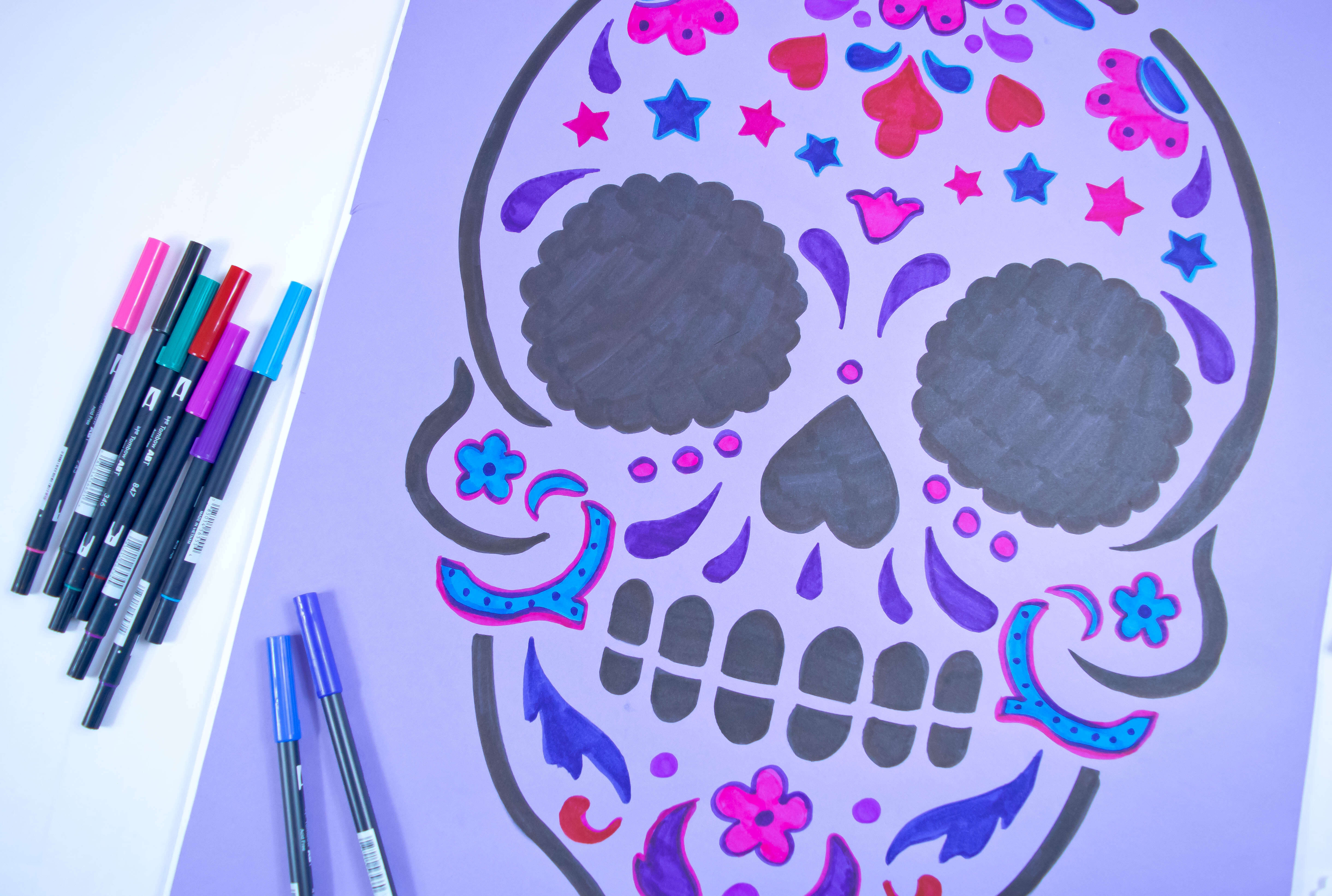 A large sugar skull is stenciled on to a purple poster board. The skull is colored in with different colored brush pens that lay next to the poster board.