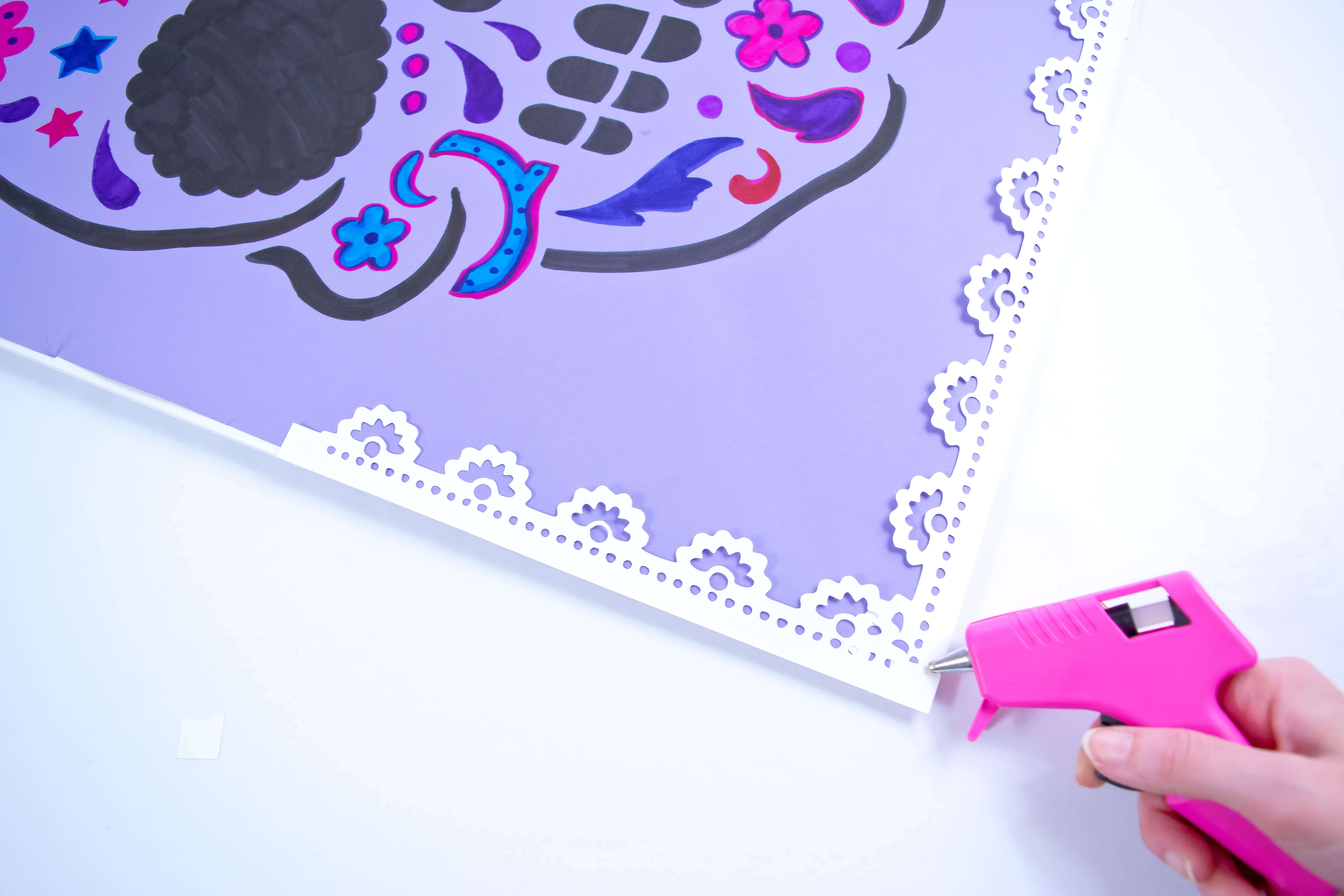 A person uses a pink crafting glue gun to attach a white paper lace border to the purple sugar skull poster.