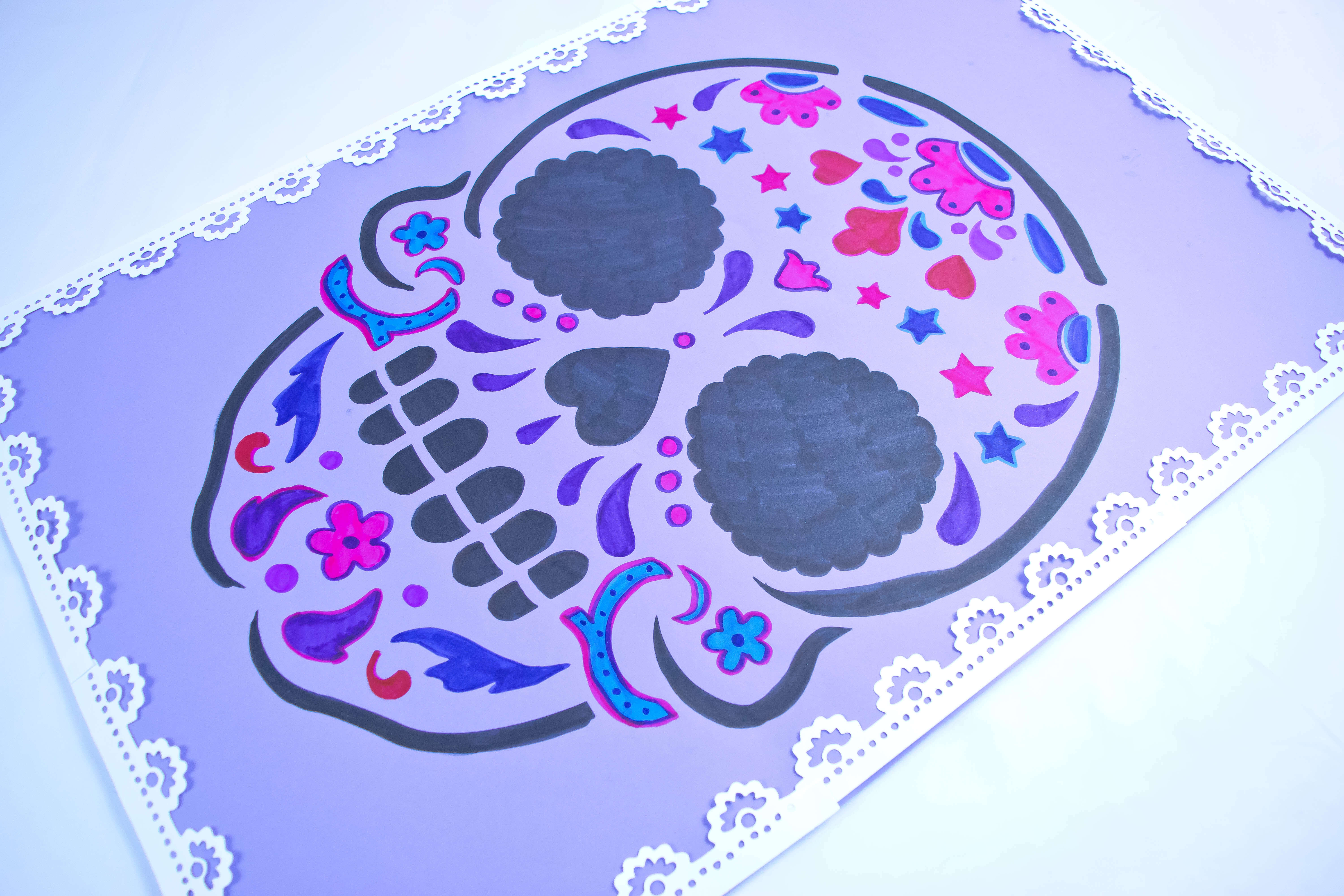 An angled view of a purple paper poster decorated with a colorful sugar skull craft. White paper lace lines the edges. 