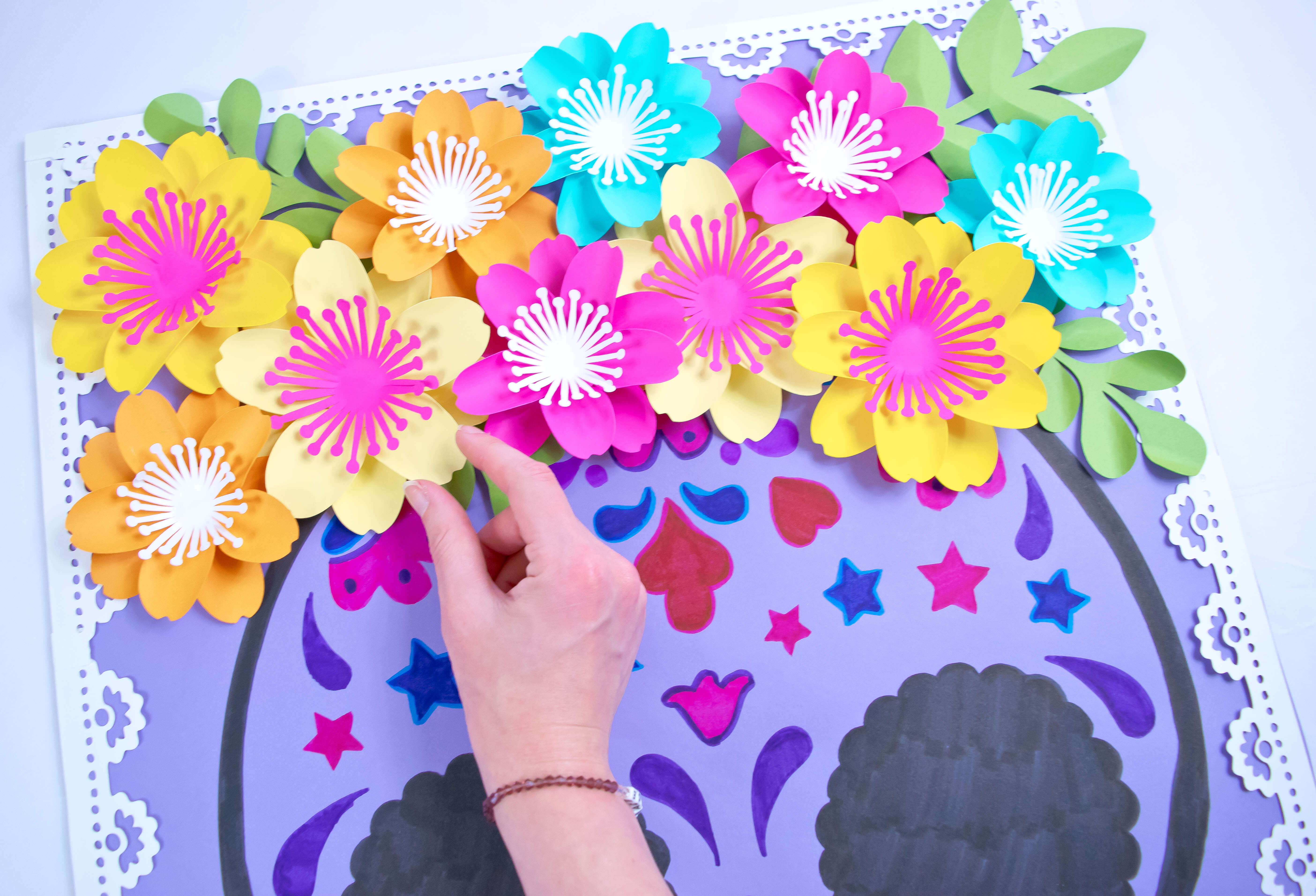 A completed Day of the Dead sugar skull drawing is complete with a cluster of colorful paper flowers in shades of orange, yellow, pink, and blue with green leaves. A woman’s hand perfectly places the last yellow flower.