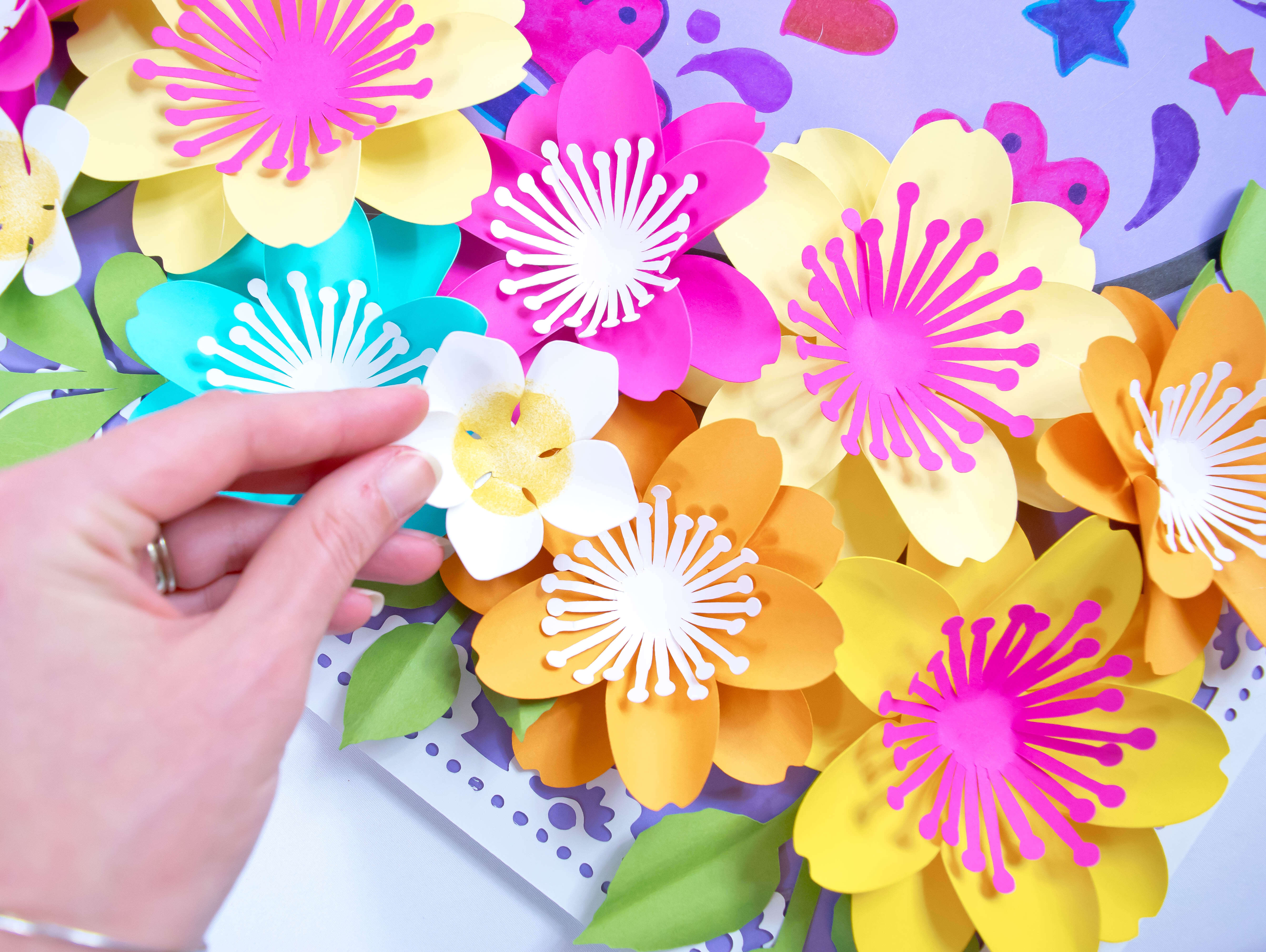 A cluster of small, colorful paper flowers in shades of orange, yellow, blue, and pink with white and pink centers and green leaves. A woman’s hand places one small white flower into the cluster.