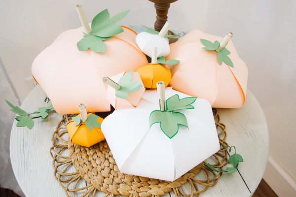 A pile of orange and white paper pumpkins with green leaves sit atop a rattan placemat. 