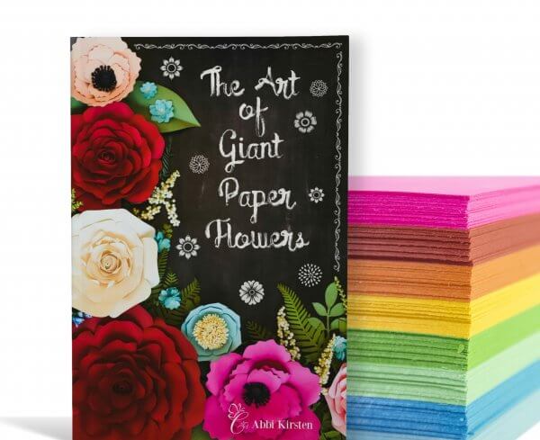 The Art of Giant Paper Flowers Book, with a black cover and red, pink, peach, and light blue roses and other flowers. Behind the book is a stack of cardstock in a rainbow line. 