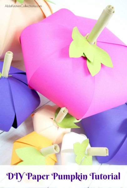 Brightly colored paper pumpkins along with muted colored paper pumpkins are piled on a white wooden table.  