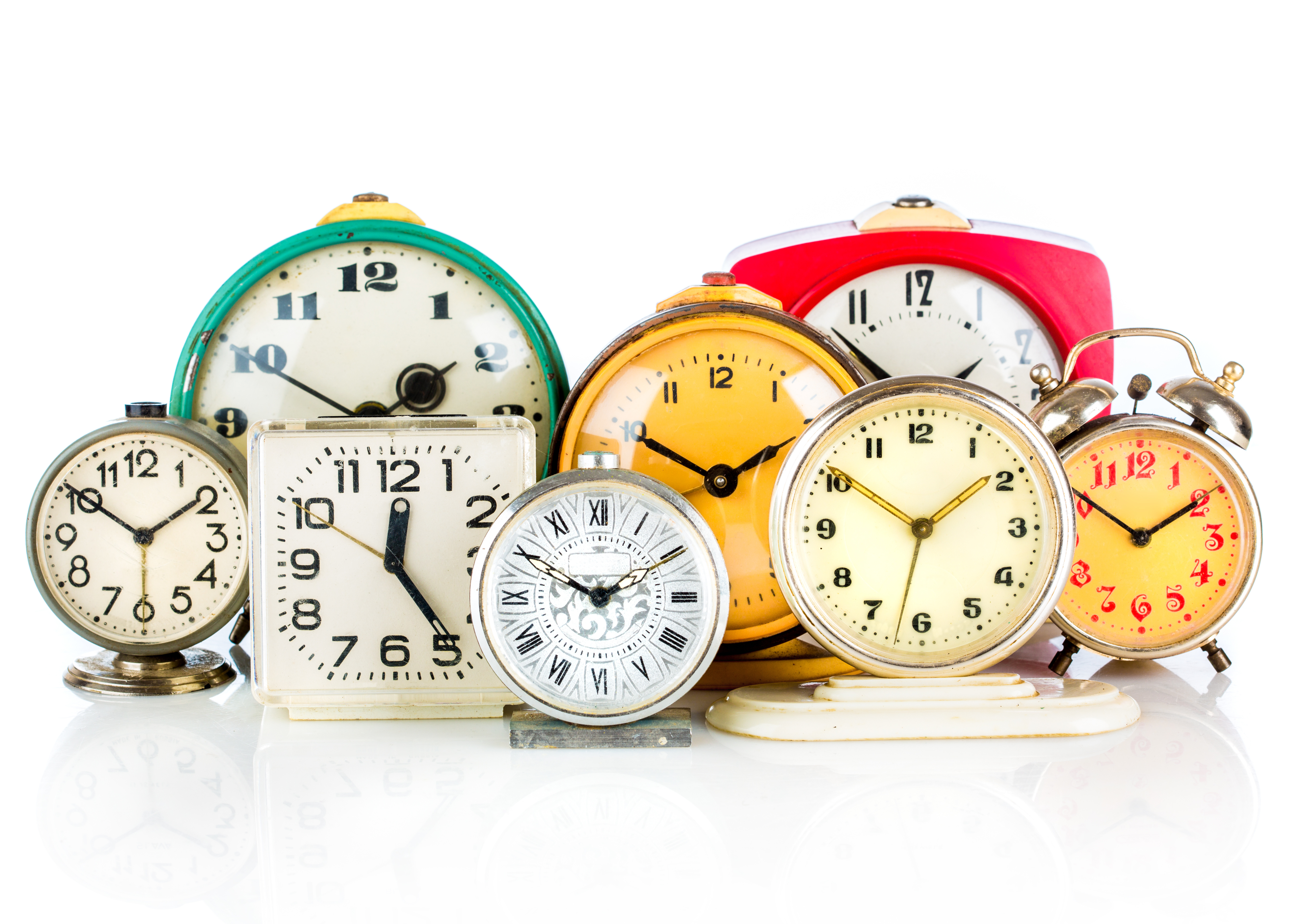 Several different analog clocks, all set to different times, are gathered together on a white table with a white background. 
