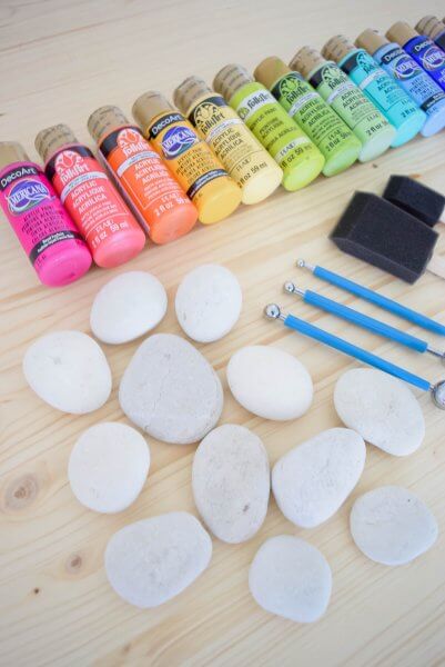 A group of rock painting supplies on a wooden table including paints, white rocks, and paintbrushes. 