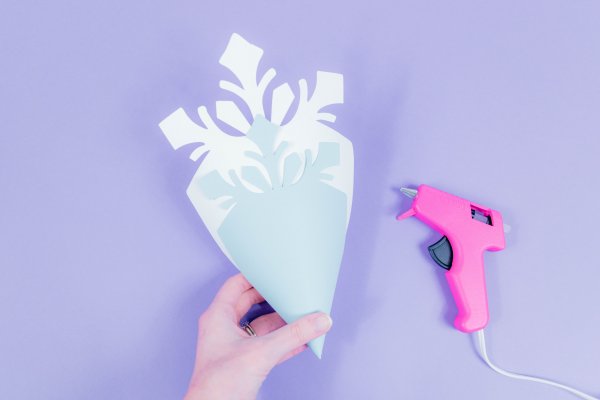 Abbi demonstrates how to stack and glue together the snowflake petals. A large light blue snowflake petal is stacked into a medium sized, darker blue petal. A pink glue gun sits on the table to the right.