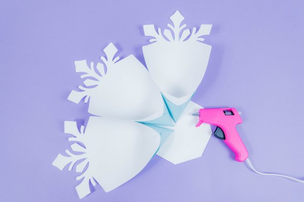 Three snowflake petals are glued to the 8-sided base. A pink glue gun sits on the table next to the in-progress paper snowflake.