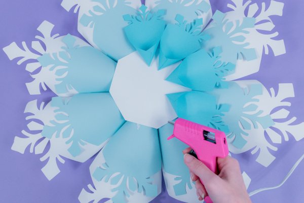 Abbi continues to glue four small snowflake petals to the front side of the giant paper snowflake using a hot glue gun.