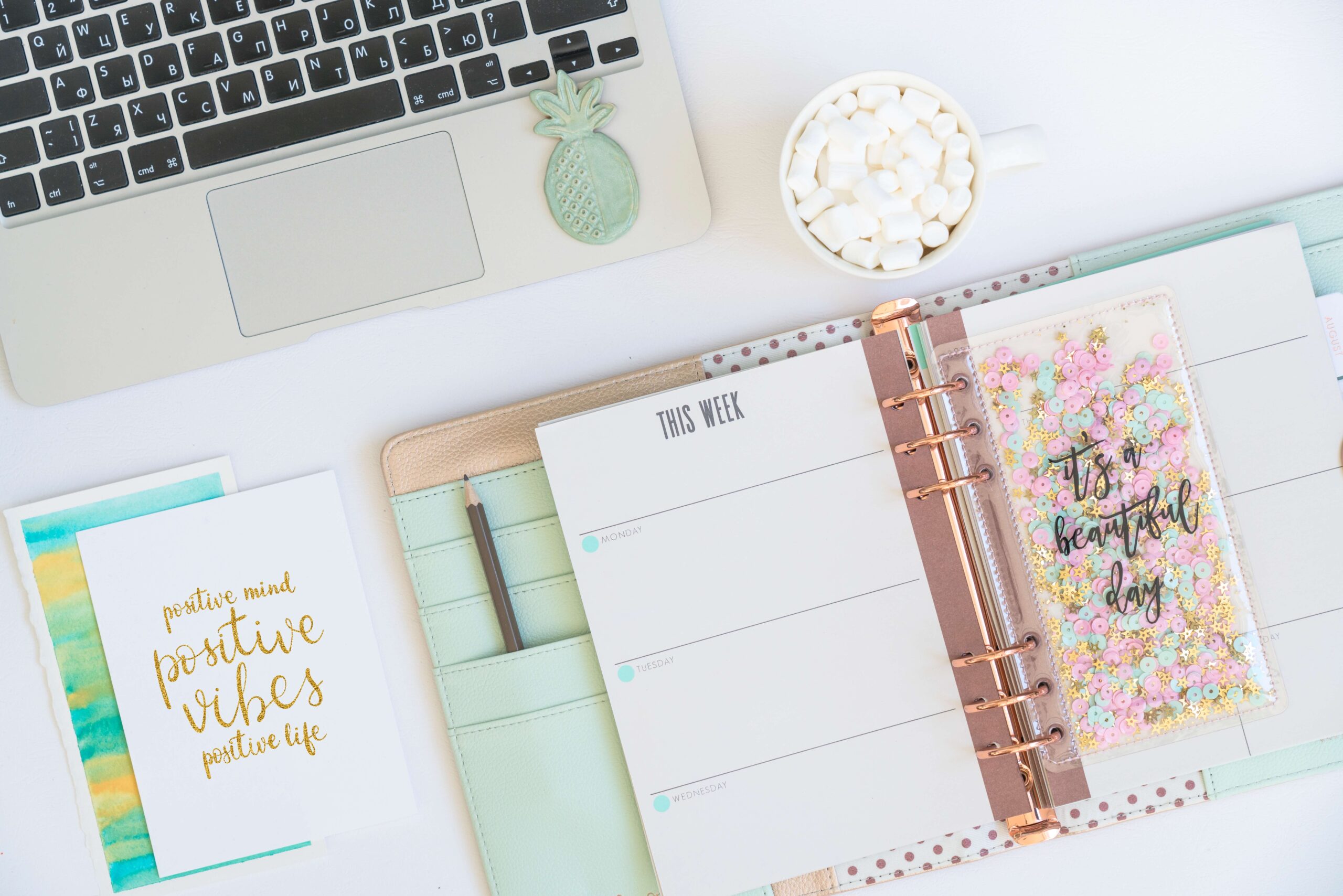 An overhead image of an open planner on a white desk, a laptop, a cup of coffee topped with marshmallows, and a card that reads "Positive mind, positive vibes, positive life."