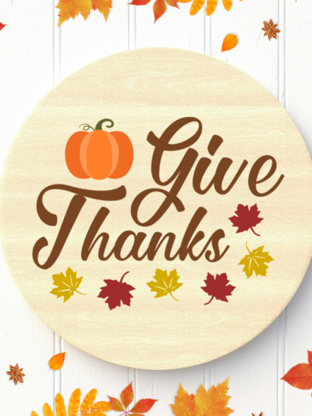 A circular piece of light wood with the words "Give Thanks" in a nice script font surrounded by a pumpkin and autumn leaves. The wood circle lays on a white wooden table surface with fall leaves at the bottom of the craft. 
