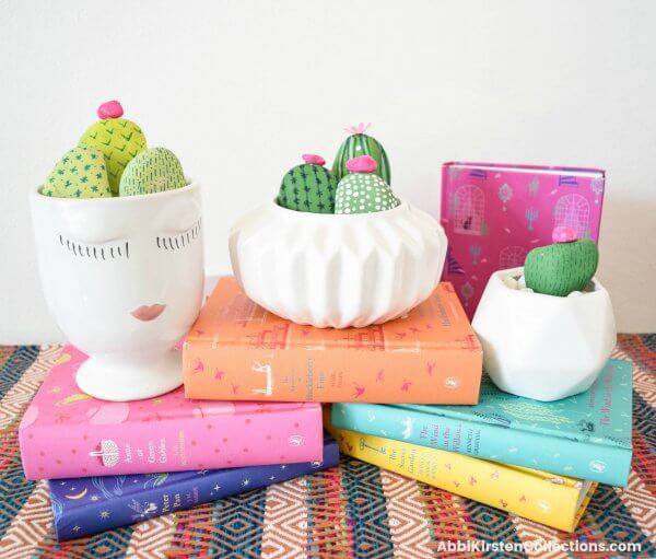 Cactus painted rocks arranged in white planters set atop colorful books. 