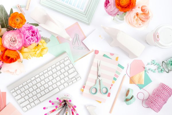 A bright but cluttered white desktop as seen from above. Scissors, flowers, a keyboard, part of a picture frame, twine, tape, and a coffee cup with pink lipstick marks are spread across the table with other craft and office supplies. 