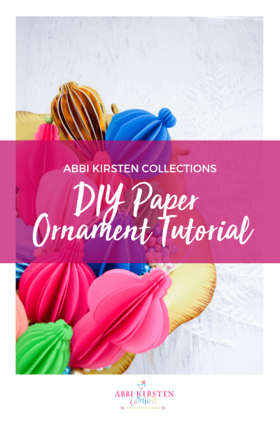 Multicolored folded paper ornaments sit in a bowl. Text over the top reads "Abbi Kirsten Collections DIY Paper Ornament Tutorial." Get your home holiday ready with these easy-to-use templates!