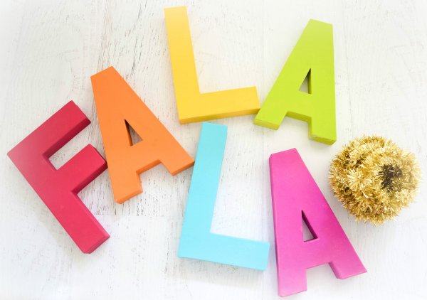Fa La La letters and rolled up gold tinsel sit on a white surface. The decorative Christmas tree letters are painted in bold bright colors. 