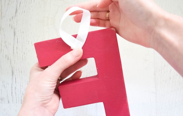 Abbi Kirsten's hands show how to attach a loop of white ribbon behind a red letter "F" so the letters can hang on the Christmas tree or anywhere you want to add decorative sayings. 
