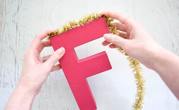Abbi Kirsten's hand hold gold tinsel tightly around the sharp corners of the red letter "F."