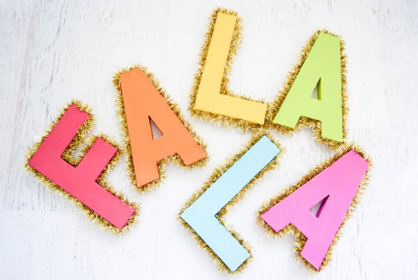 Brightly colored, bold, and gold tinsel-trimmed letters spelling out the Christmas tune "Fa La La" lay on a white wooden surface. You can make any cardboard letter into any event decor. 