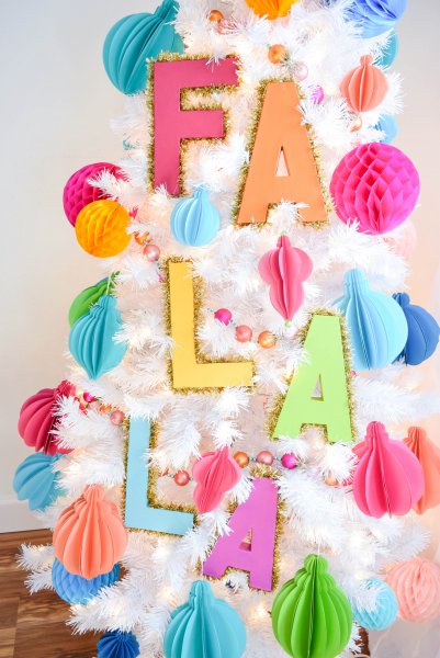 A white Christmas tree with colored paper balls and the words "Fa La La" hung vertically down the tree. They are decorative tinsel Christmas tree letters. 