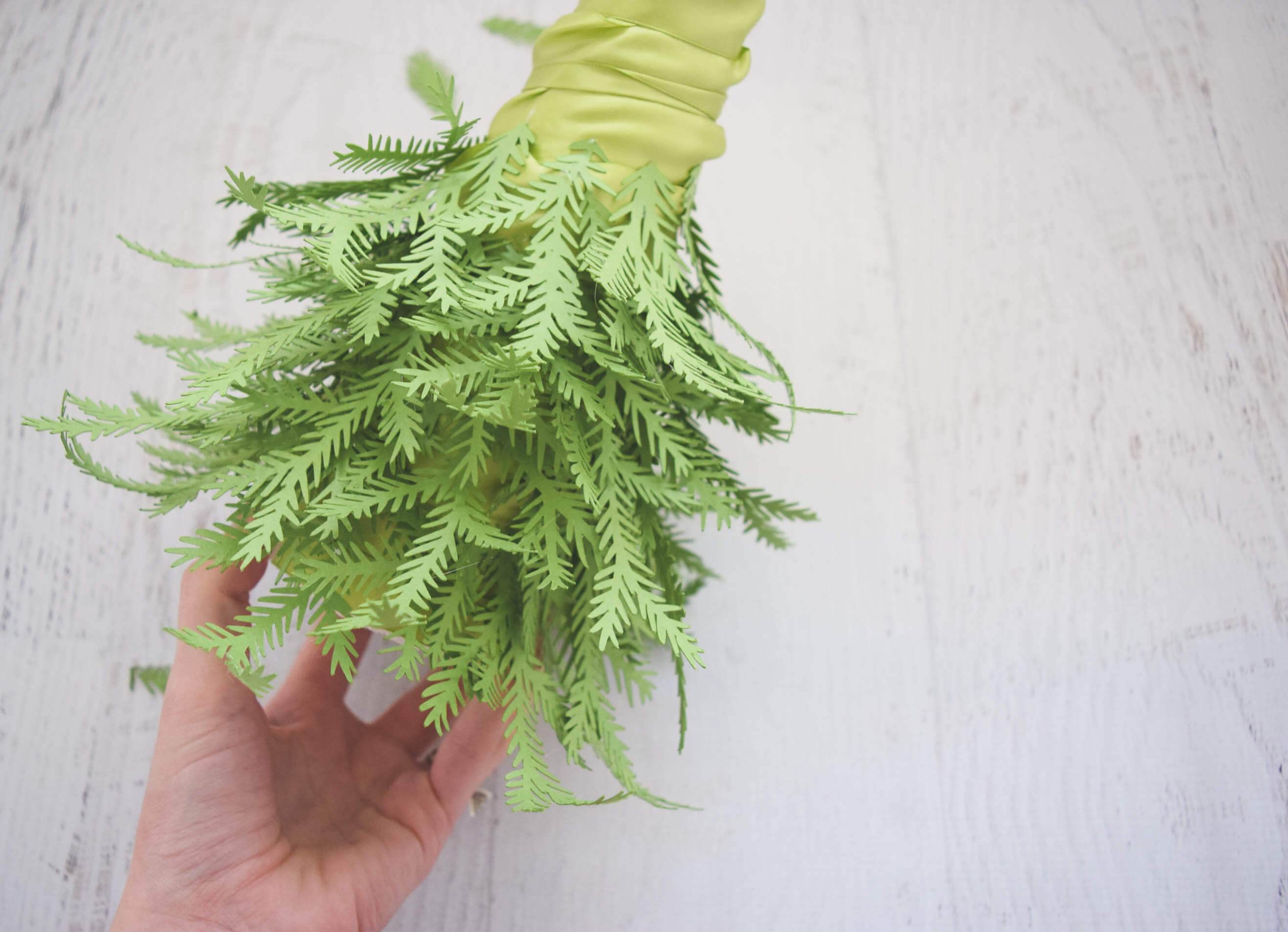 A woman's hand holds the in-progress paper Christmas tree, half-way filled with green paper pine leaf branches.