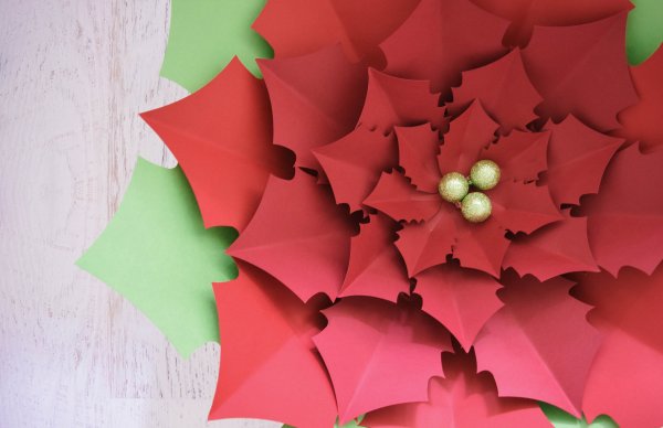 A completed giant Noel Poinsettia paper flower with red petals and green leaves, completed with a gold holly berry center.