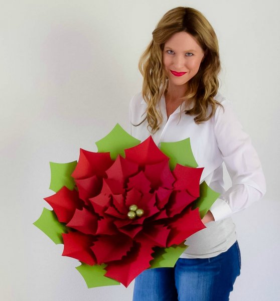 Abbi Kirsten holds the giant Noel Poinsettia flower in her hands. She's wearing jean and a white long sleeve shirt, and smiling at the camera.