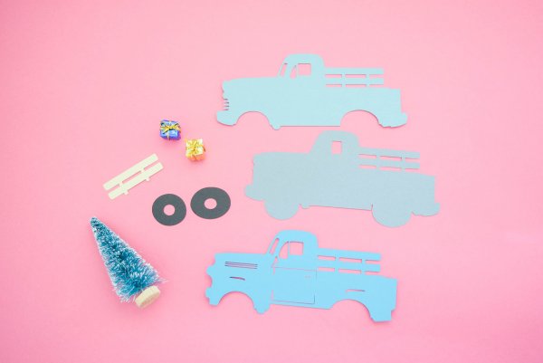 On a pink backdrop lay truck pieces in shades of blue paper, black tires, small presents and a mini evergreen are all the parts needed to make a vintage Christmas truck ornament. 